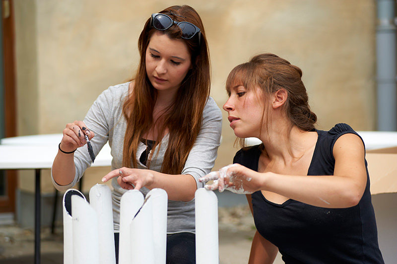 Two young females working on an architect project using their hands and a bursg. One hand covered in white paint.