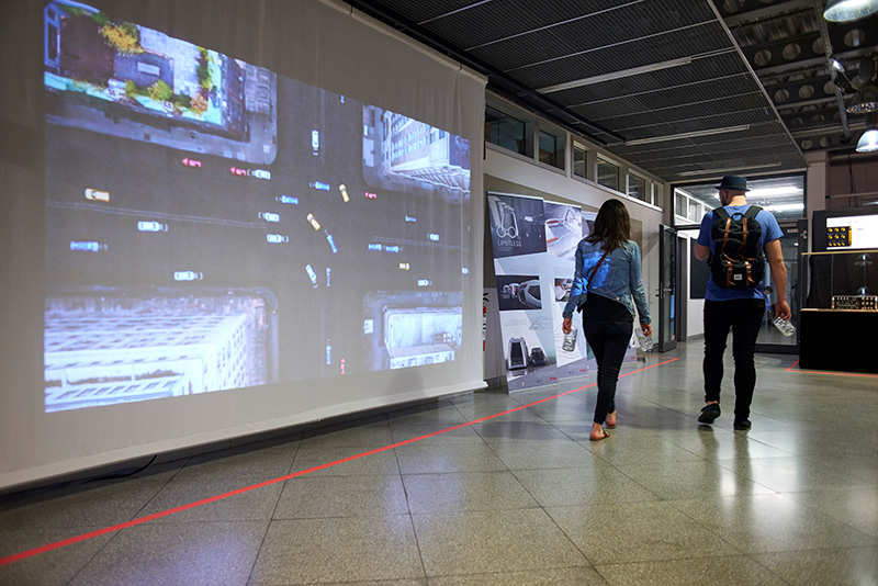 Two students walking past a screen that is being projected on. The projection shows an aerial view of an urban intersection